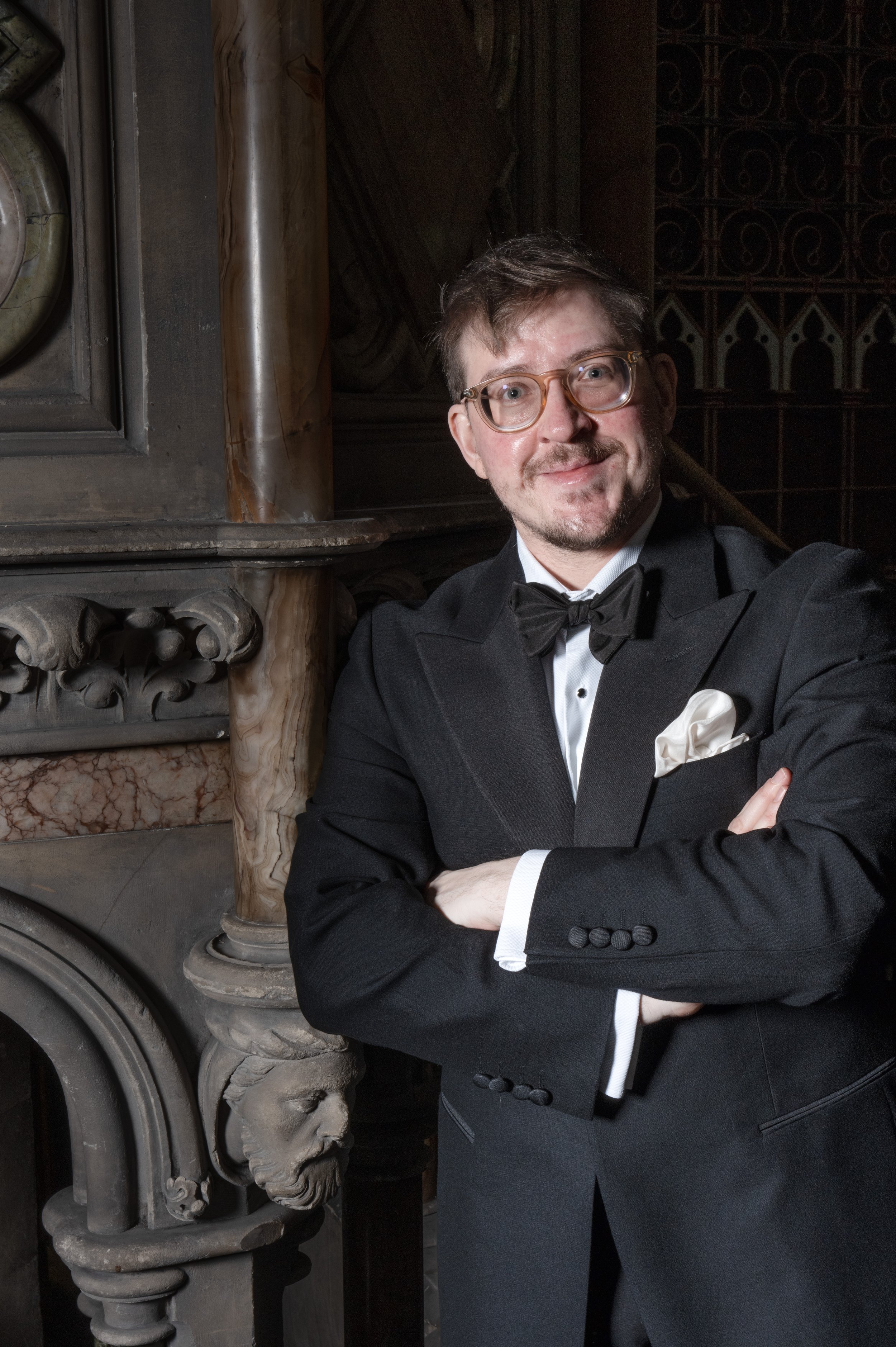 A man in a black tuxedo with a bow tie and white pocket square, wearing glasses, standing with his arms crossed in front of an ornate stone fireplace with a carved lion's head and decorative details.