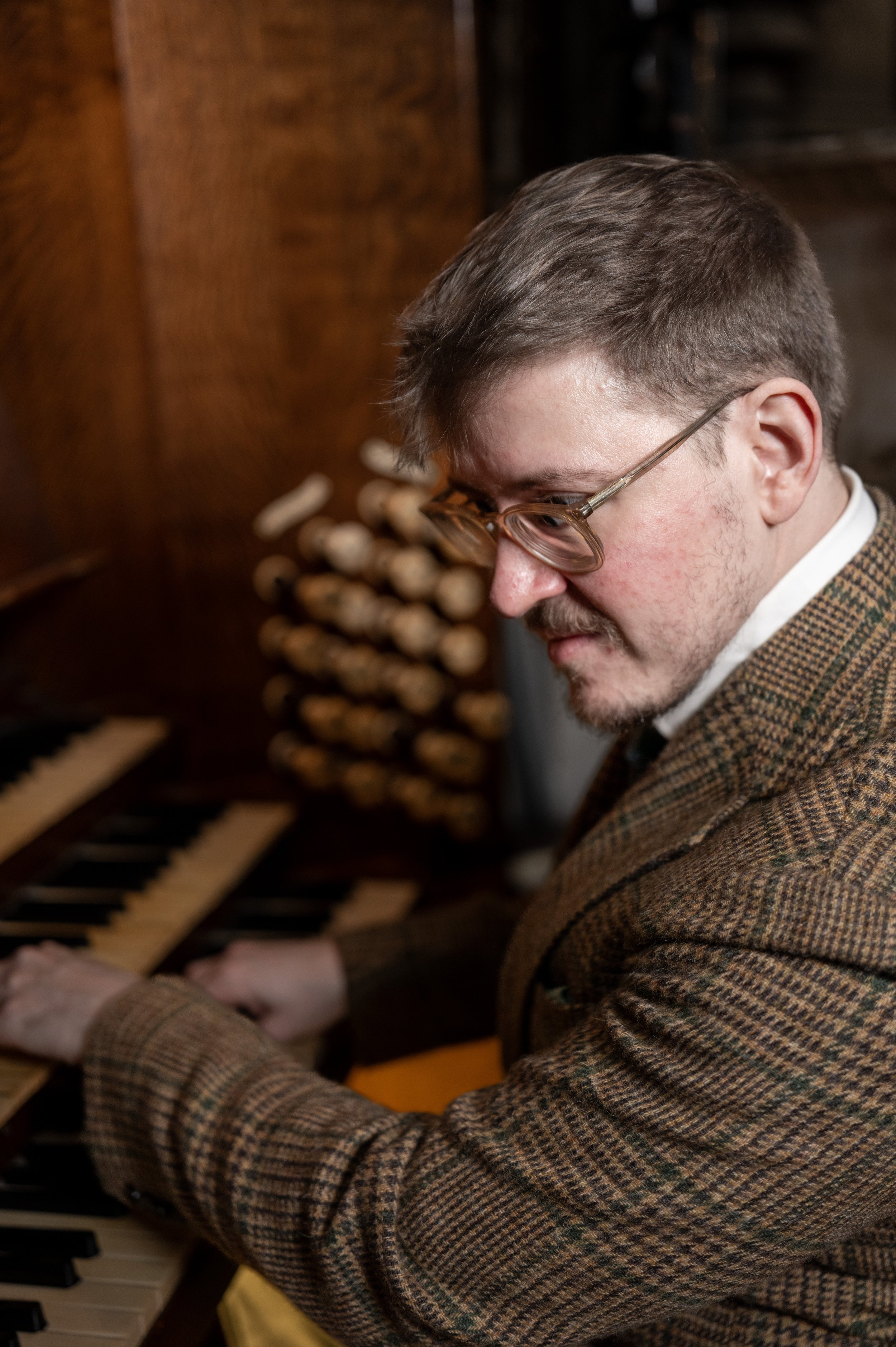 A man with glasses, wearing a plaid jacket, playing a piano.