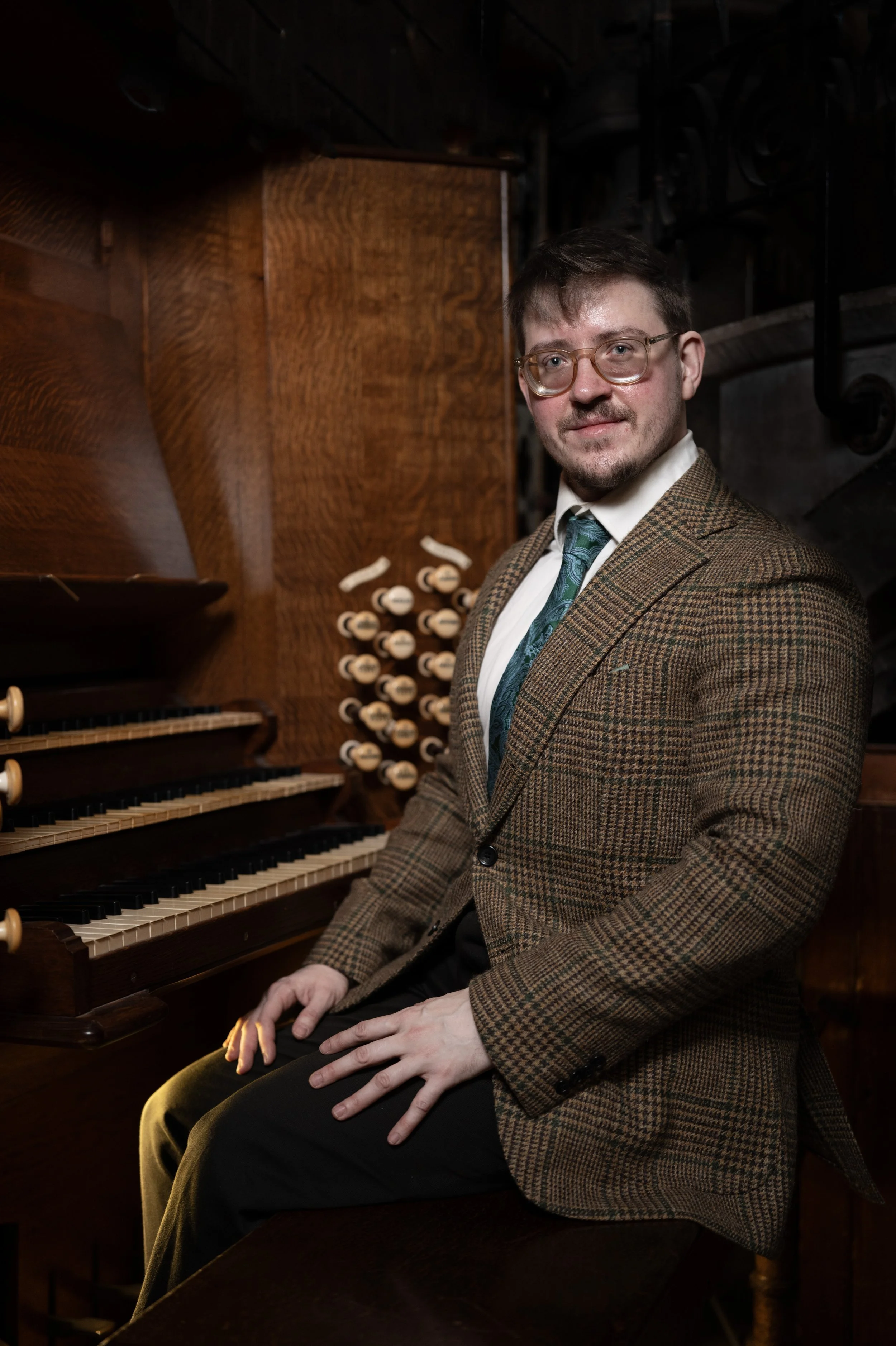A man wearing glasses and a plaid blazer sitting at a pipe organ, with a wooden background.