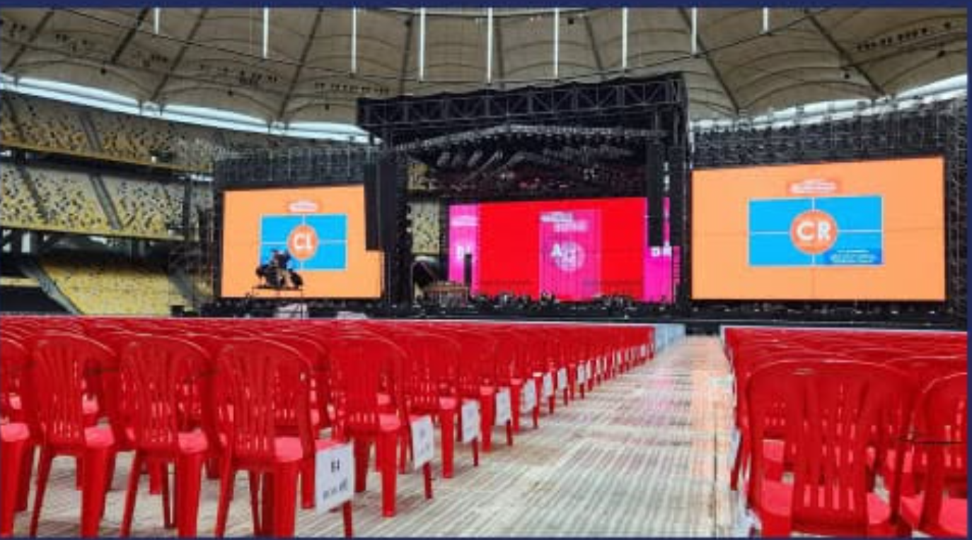 Empty seats with red chairs and numbered signs, setup in front of a large stage with multiple screens at a stadium or arena.
