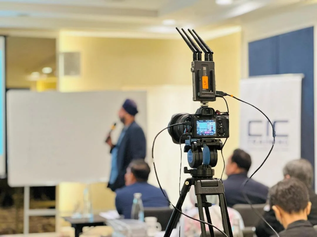A camera on a tripod capturing a speaker at a conference, with an audience seated in front.