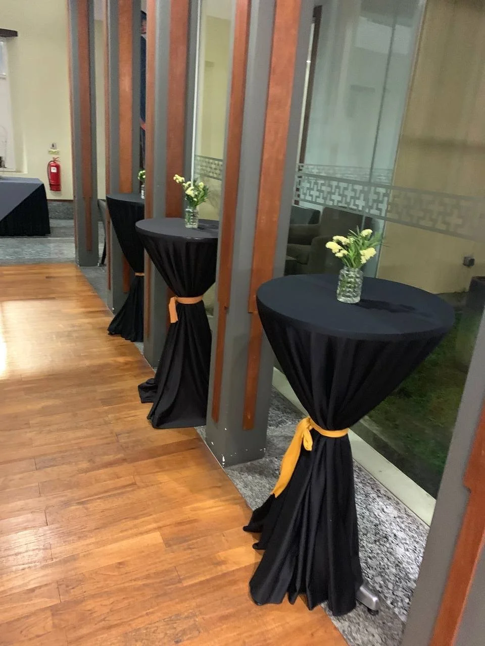 Three standing tables with black tablecloths and yellow tied sashes, each with a glass vase containing white flowers, positioned along a wall with glass window panels in an indoor setting.
