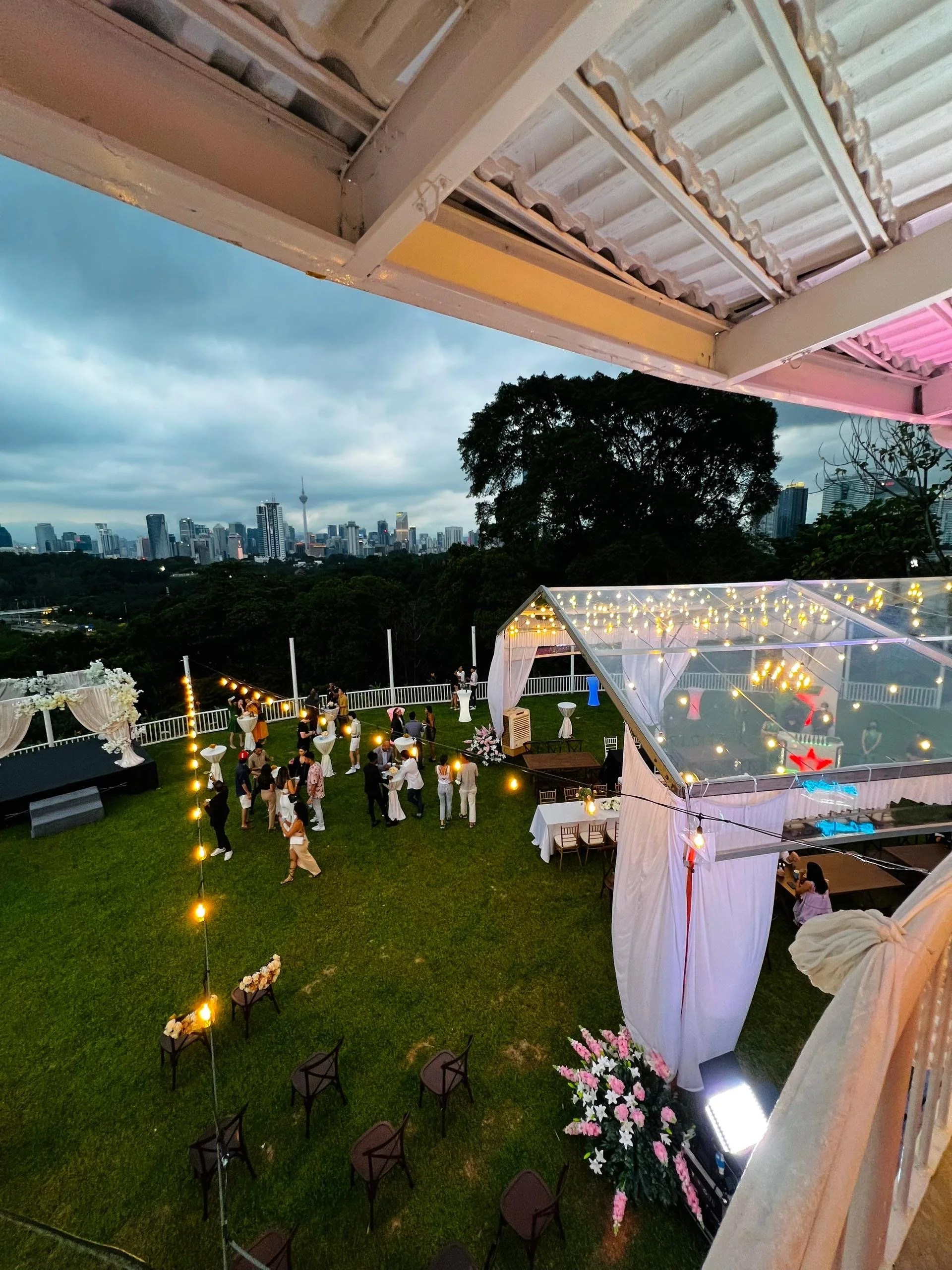 Outdoor celebration or wedding reception with string lights, tables, and people gathered on a grassy area, skyline cityscape in the background during dusk.