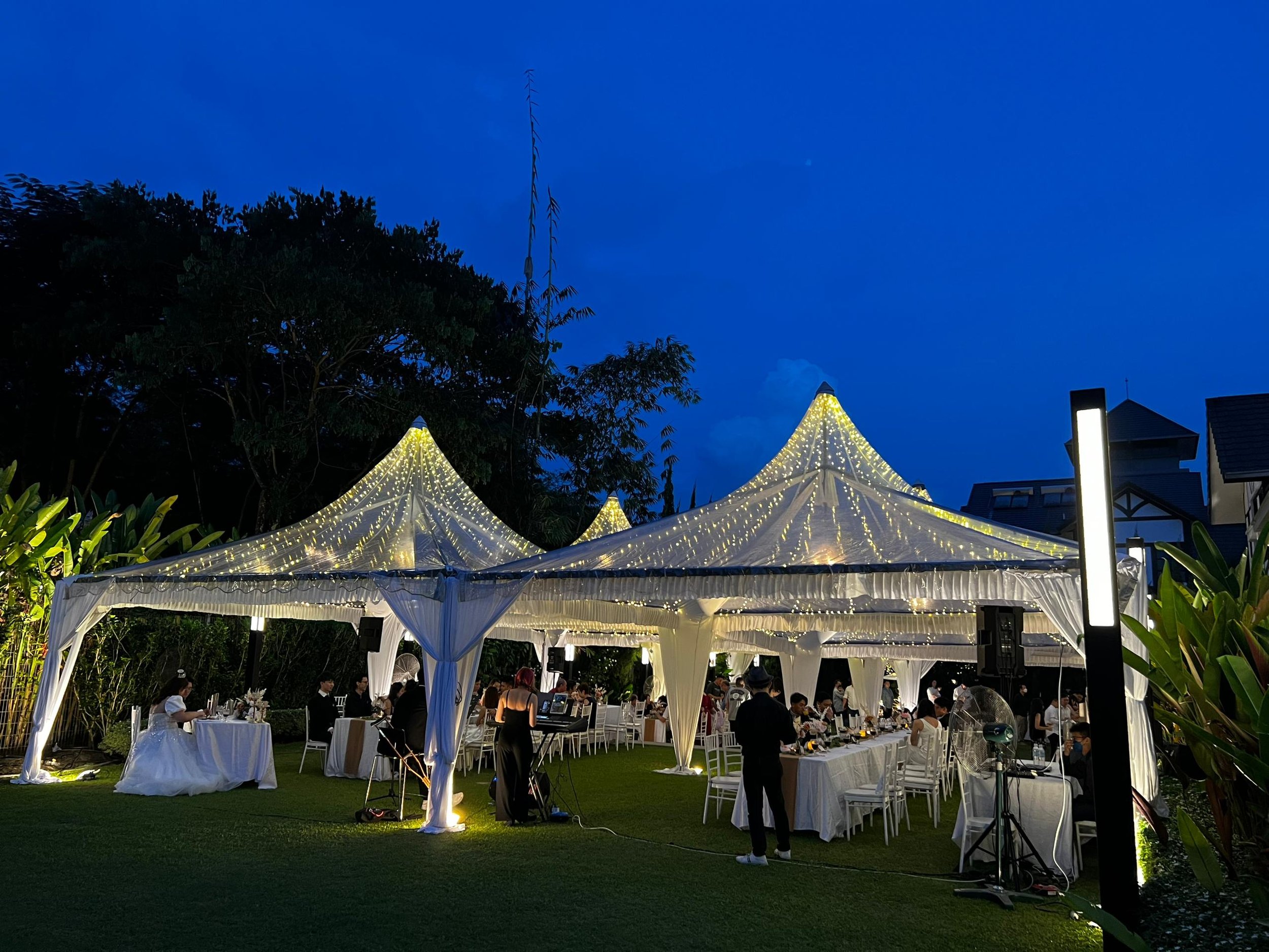 An outdoor evening event under white tents decorated with string lights, with people dining at tables, surrounded by greenery and lit by lanterns.