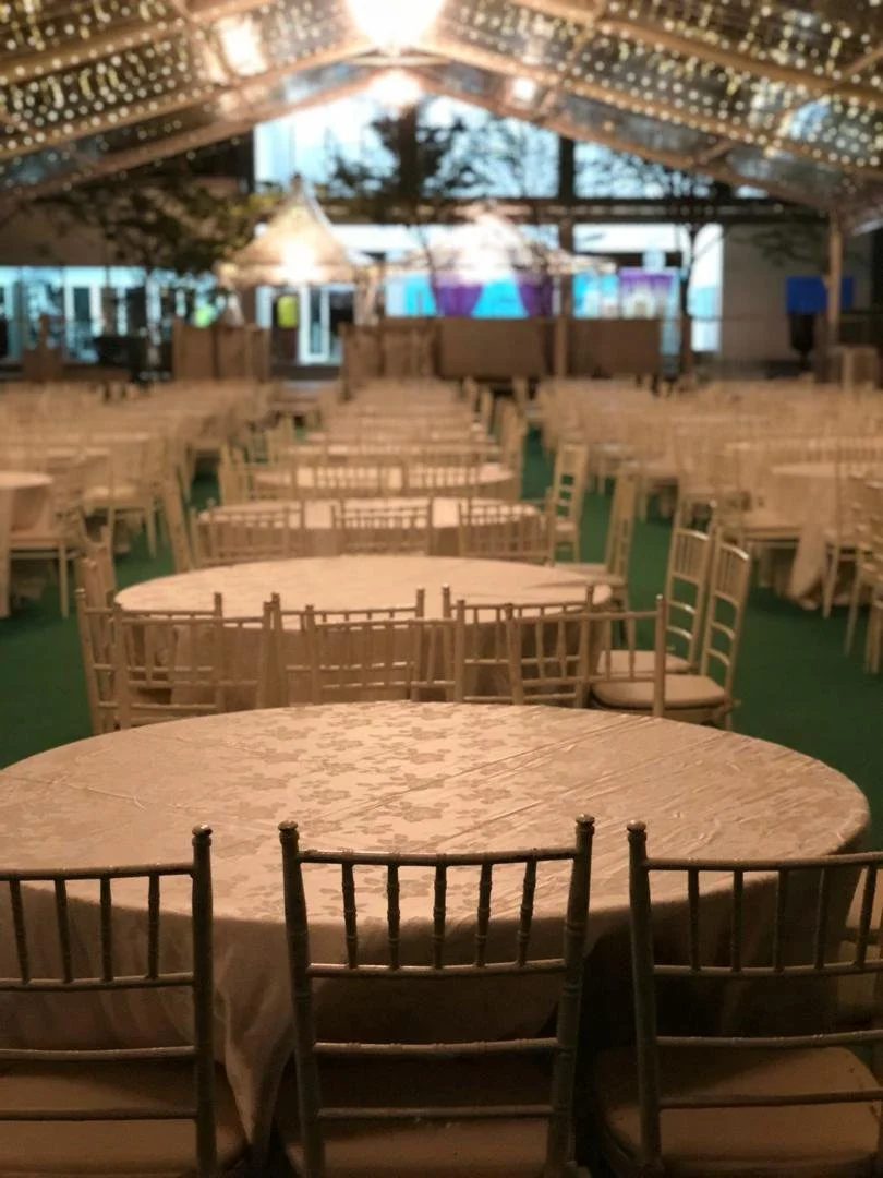 Empty round tables with beige tablecloths and wooden chairs in a decorated event tent with string lights.