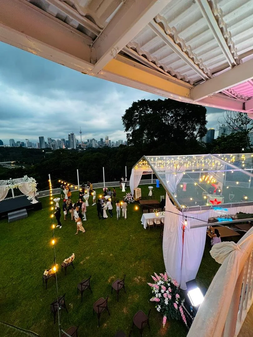 An outdoor celebration on a rooftop with city skyline in the background, decorated with string lights, flowers, and a transparent tent, where people are gathering and socializing.