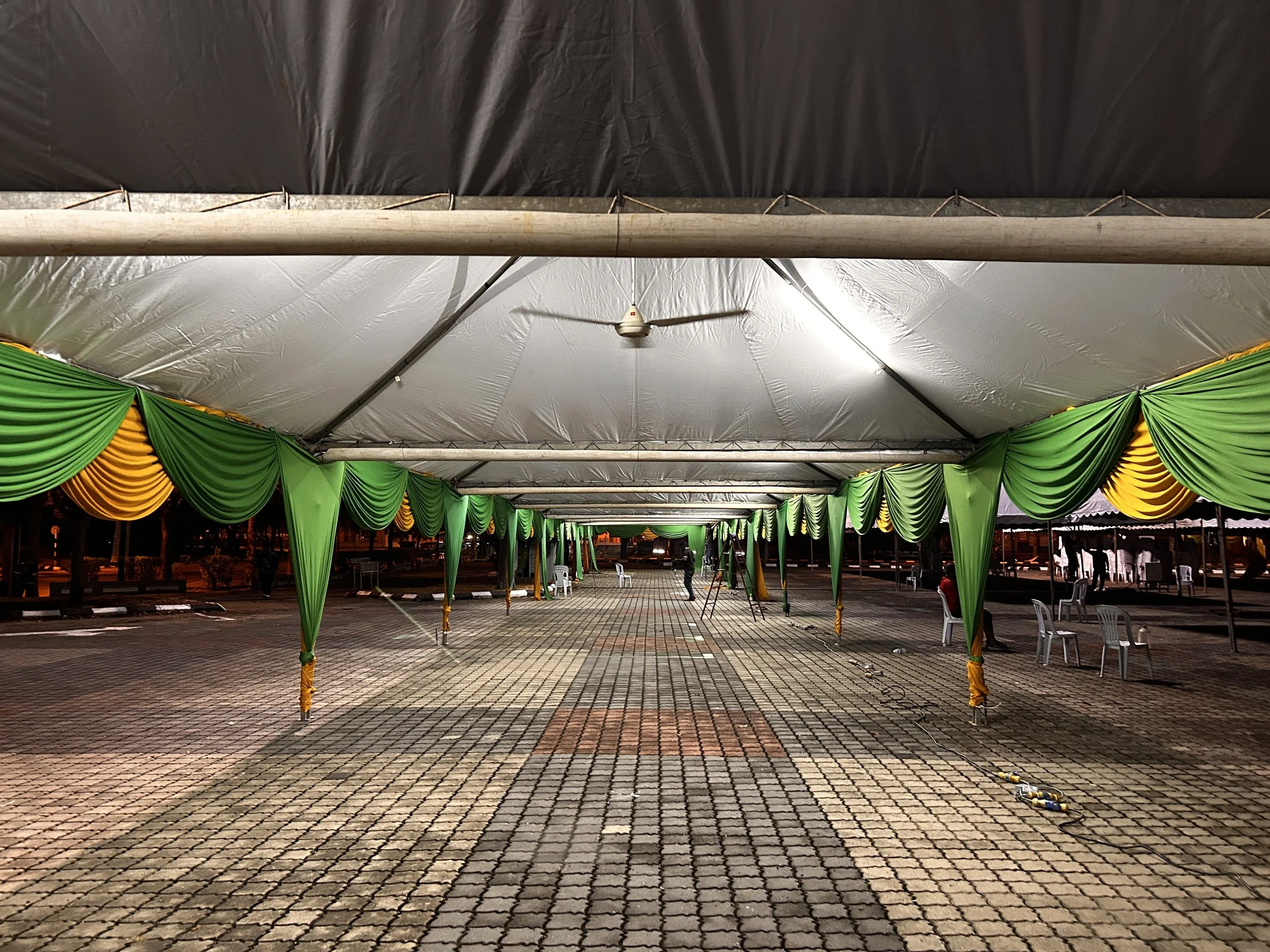 A large outdoor event tent with green and yellow draped fabric, empty tables and chairs, set up on a paved area at night under bright lights with ceiling fans, with some equipment and people visible in the background.