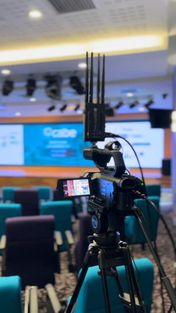 Video camera on tripod recording a presentation in a conference room with rows of chairs and a large screen at the front.
