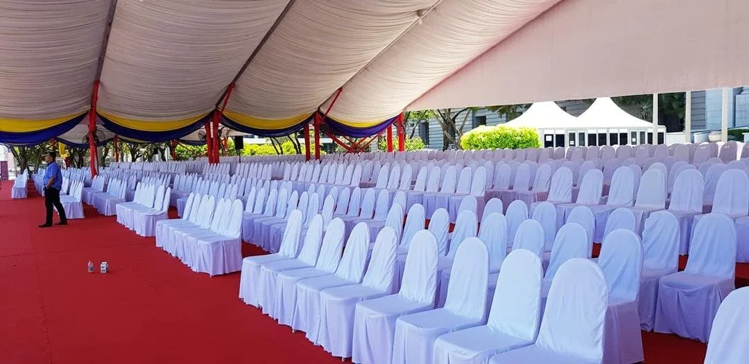 Empty white chairs arranged in rows for an outdoor event under a large tent with red, blue, and yellow accents.