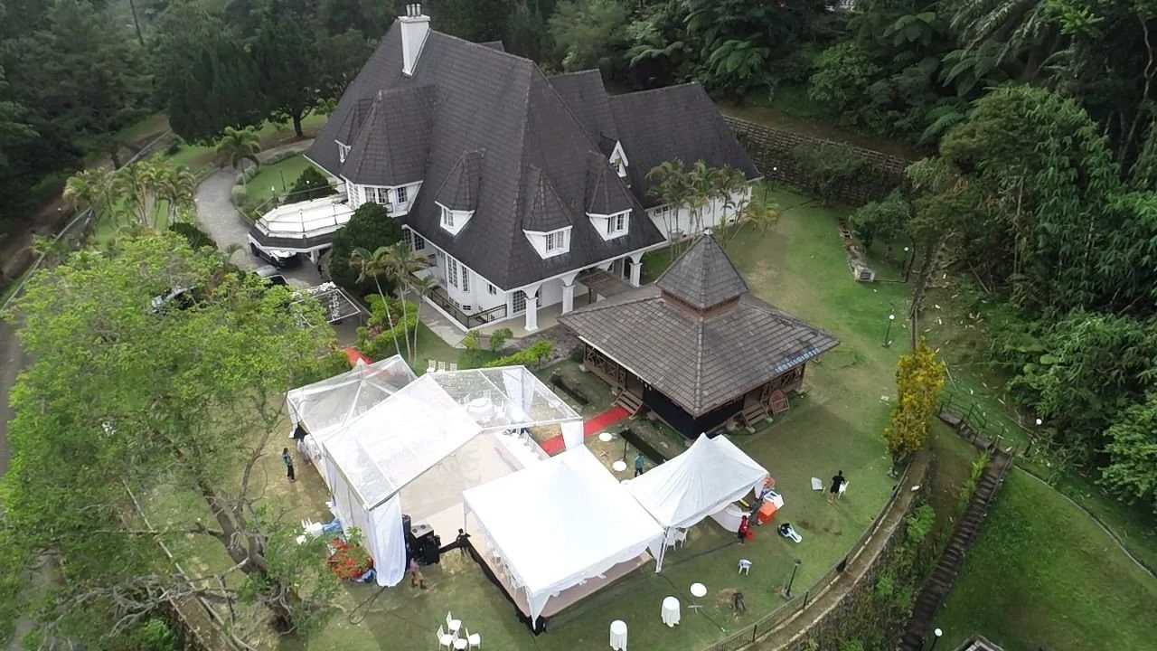 An aerial view of a large estate with a dark gray house, a gazebo, and multiple white tents set up on the lawn, surrounded by trees and greenery.