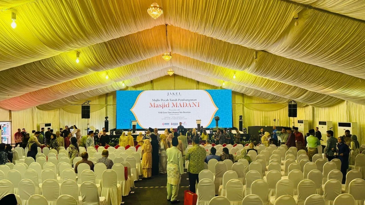 Event hall decorated with yellow drapes, filled with rows of white chairs, some occupied by attendees. Large stage with a blue screen displaying text in Malay and people gathered around.