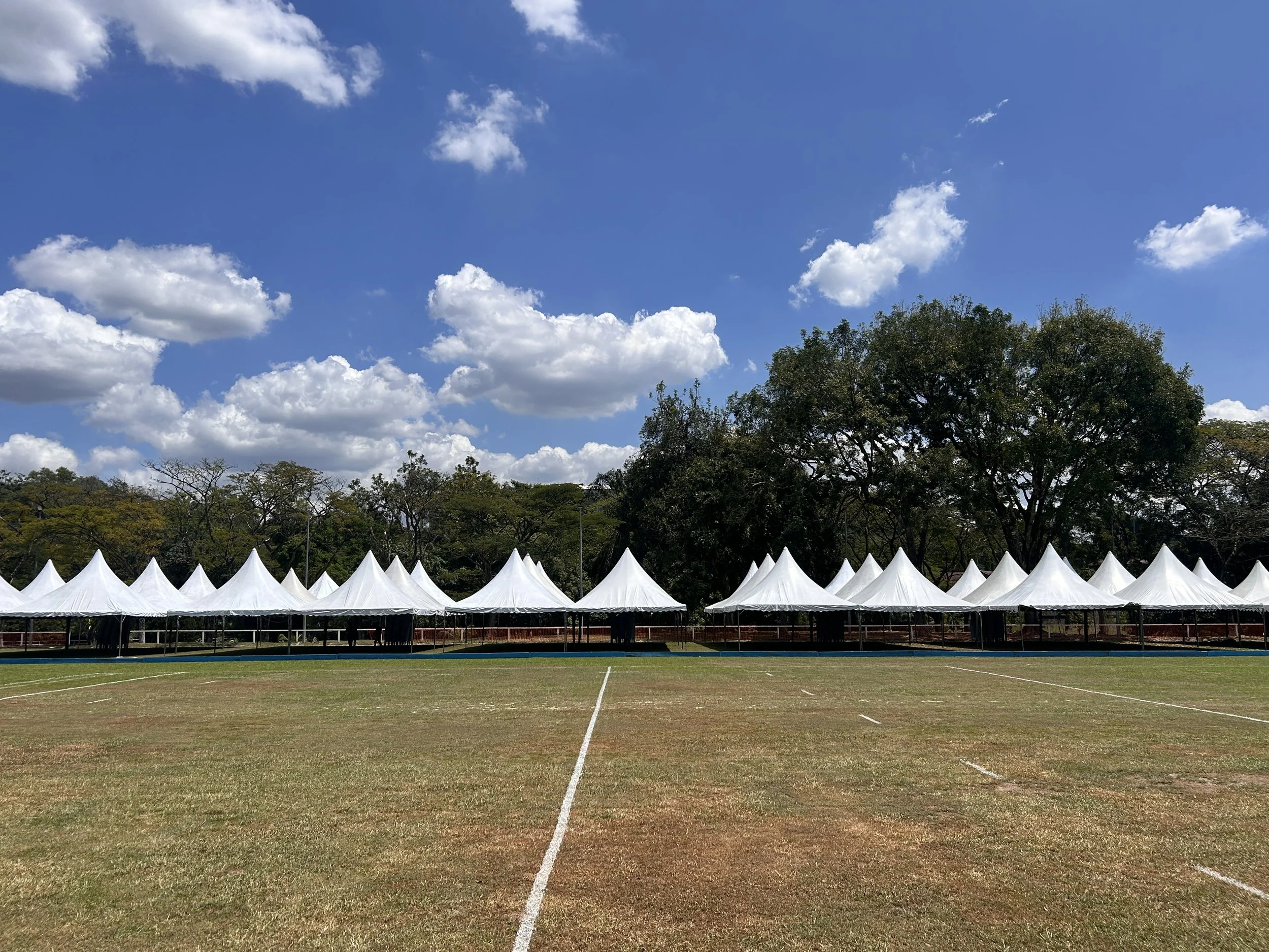 An outdoor sports field with white tents arranged in a row and trees in the background under a partly cloudy sky.