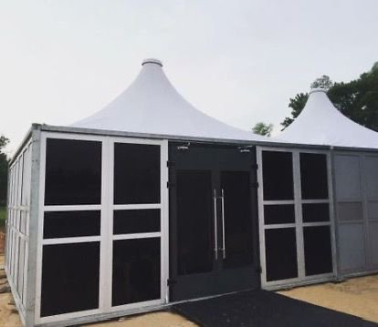 A large white tent with black panels and a black door, set up outdoors on a dirt surface with trees in the background.