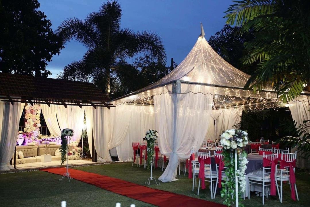 Outdoor wedding reception setup under a white canopy with string lights, decorated with flowers on stands, surrounded by tropical trees, with a red carpet walk and elegant seating arrangements.