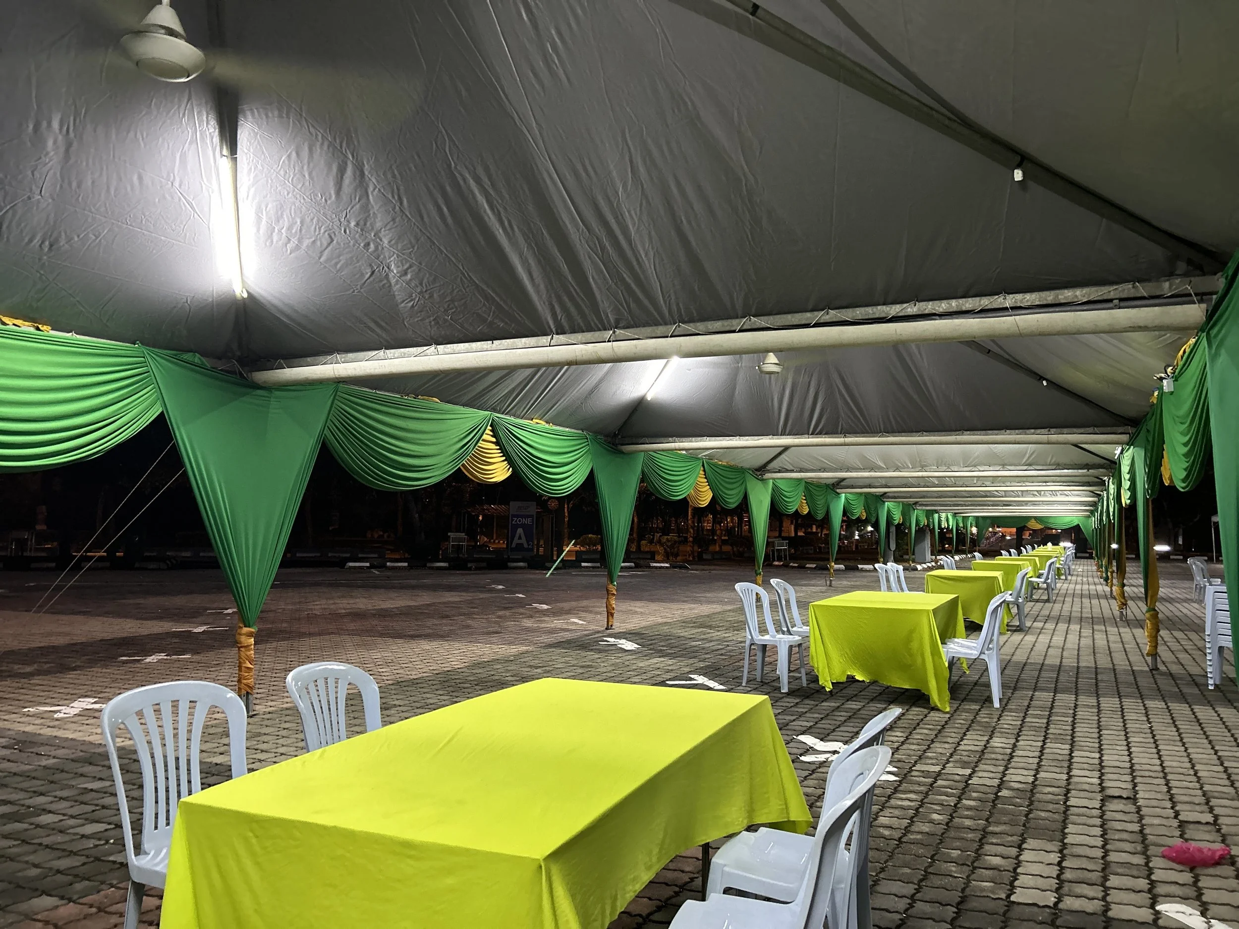Empty outdoor event space at night with yellow tables covered in bright yellow tablecloths, white plastic chairs, and a large tent with green and yellow drapes. The space is dimly lit with hanging lights.