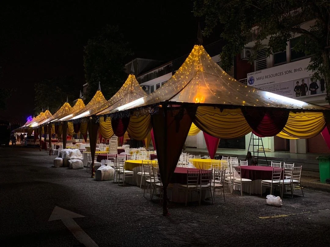 Decorated outdoor event tents with yellow and maroon drapes and string lights, set up on a street at night with chairs and tables underneath.