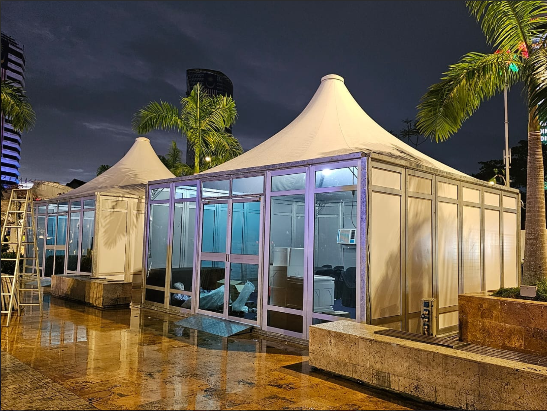 A large outdoor event tent with a pointed white canopy, surrounded by palm trees and located on a wet, reflective stone surface at night. Behind the tent, there are tall buildings and a dark, cloudy sky.