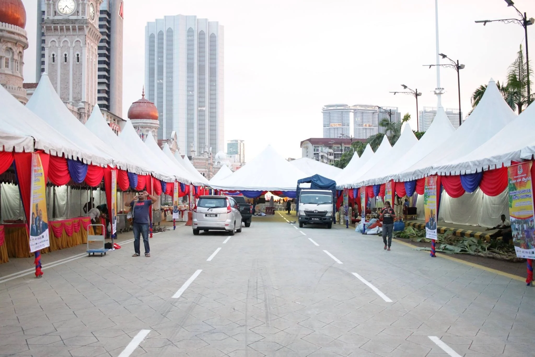 Street market with white tents decorated with red and blue drapes, set up on a paved road in an urban area with tall buildings and skyscrapers in the background.