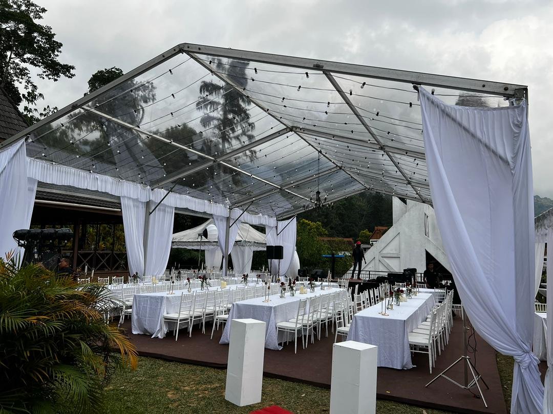 An outdoor event setup with white tables and chairs under a transparent canopy with string lights, white curtains, and a small stage area.