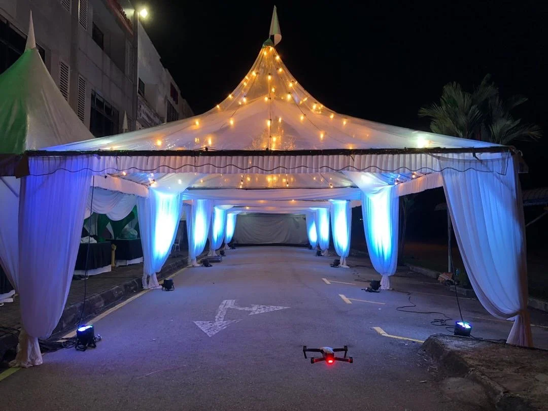 Festive outdoor event tent decorated with string lights and blue uplighting, set up on street at night, with a drone on the ground nearby.