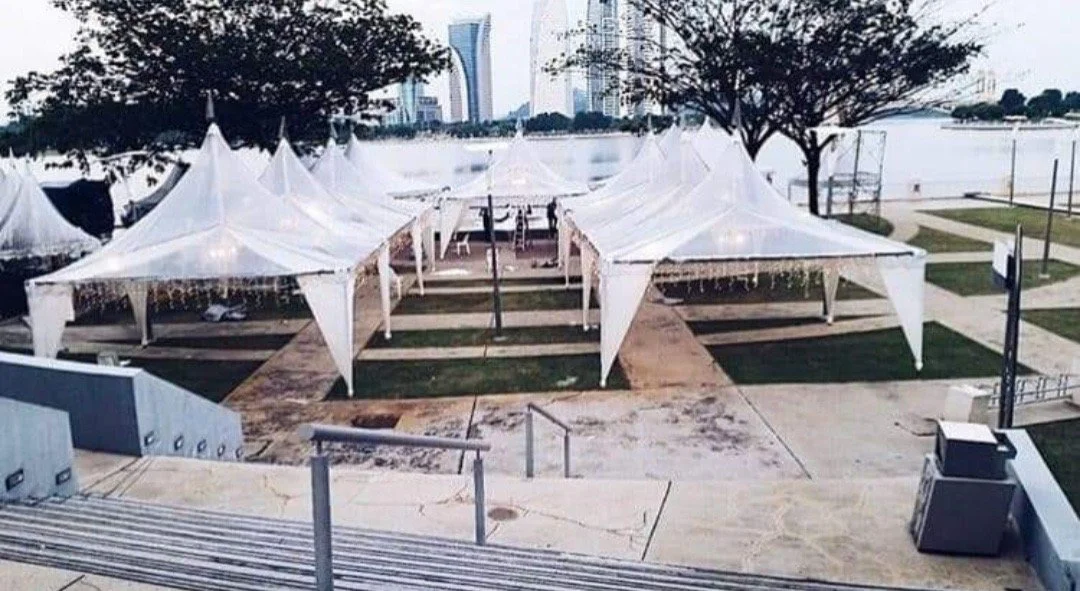 Empty outdoor event tents with decorative lights on a paved area near a river with city skyline in the background during daytime.