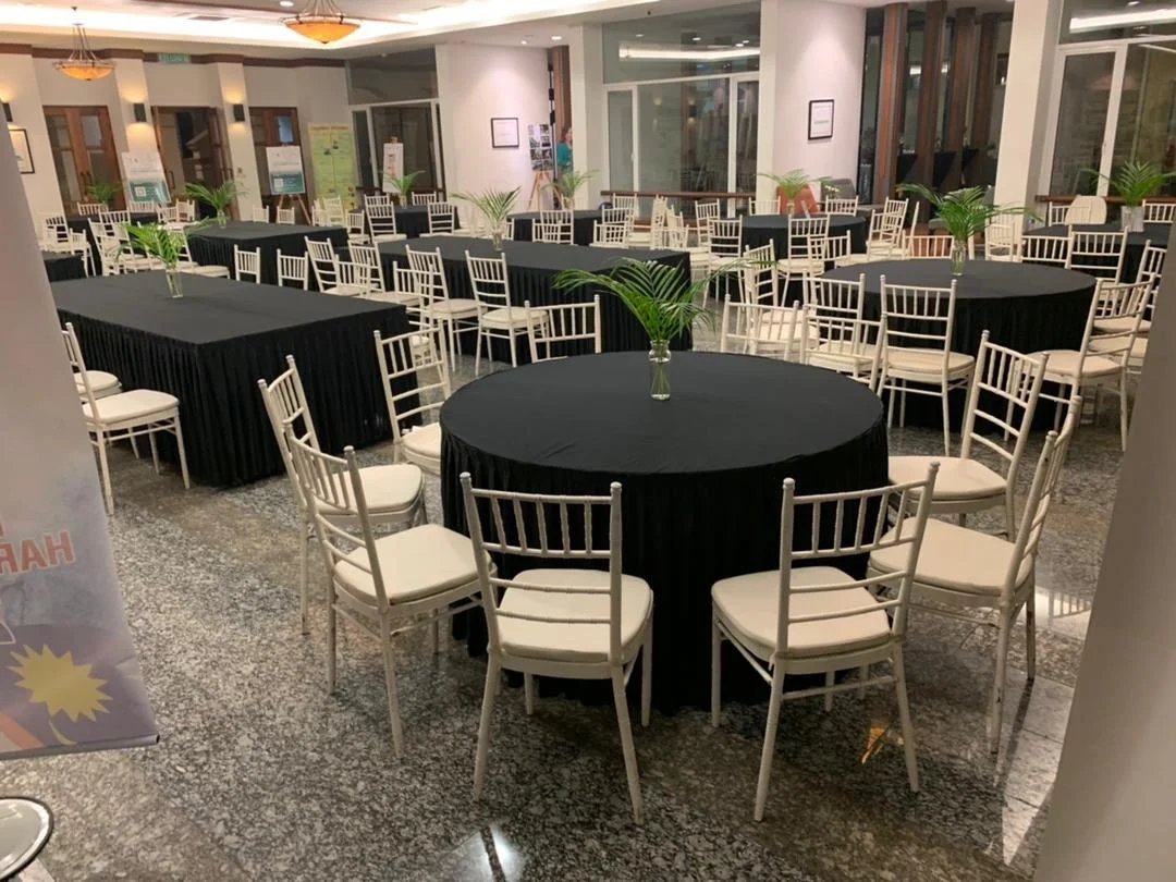 Event or banquet room with round tables covered in black tablecloths and white chairs, each table decorated with a green palm leaf centerpiece, with additional tables in the background, large windows, and some potted plants.