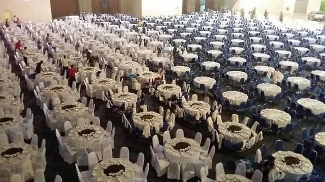 Hundreds of round banquet tables covered with white tablecloths and surrounded by white chairs, arranged in rows inside a large hall, with some people setting up or preparing the event.