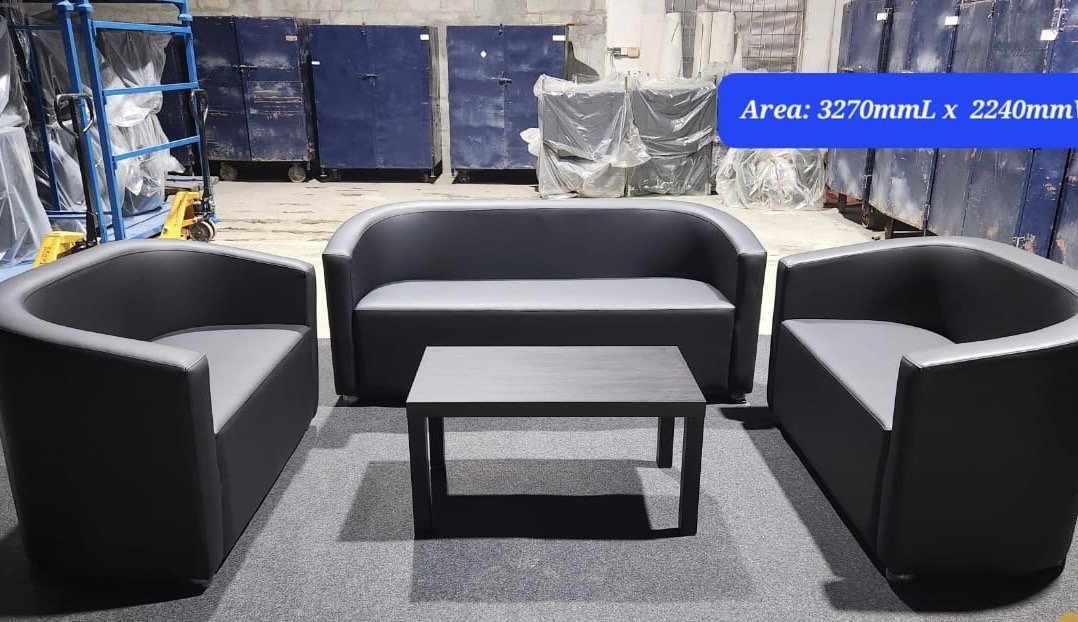 Three black sofas and a small black table on gray carpet in an industrial warehouse with blue storage bins in the background and a blue sign indicating dimensions.