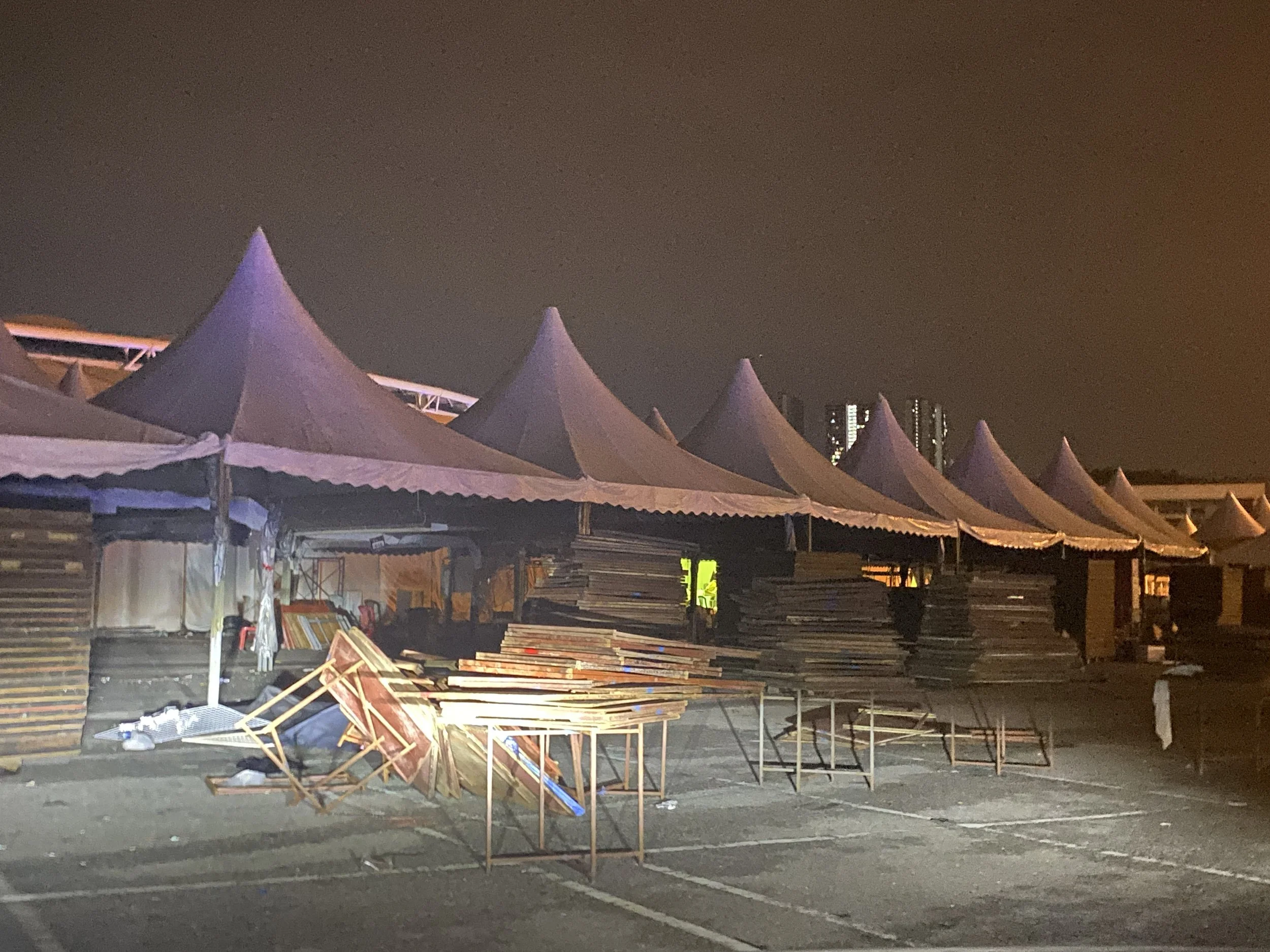 Empty outdoor market stalls with tents at night, some tables and chairs, and piles of wood, under a dark sky.