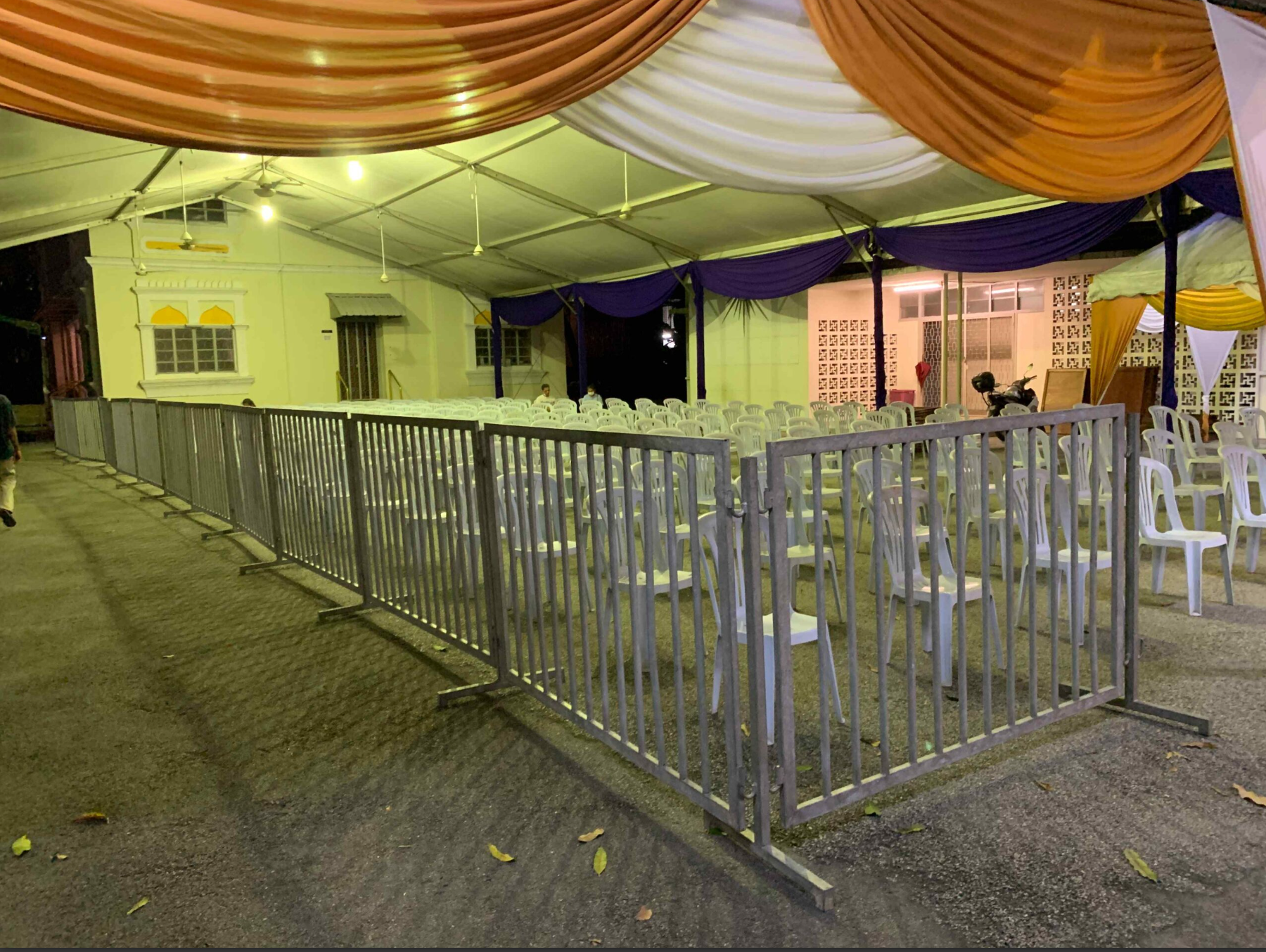 An outdoor event space set up with rows of white plastic chairs facing a stage decorated with purple and gold drapes. The area is enclosed with a metal fence, and there is a building with windows and decorative wall panels in the background. The even