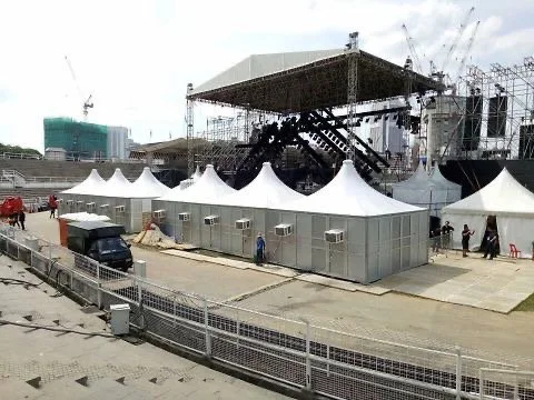 A concert stage with black curtains and scaffolding, surrounded by white tents and temporary structures, with a few trucks and workers nearby, under an overcast sky.