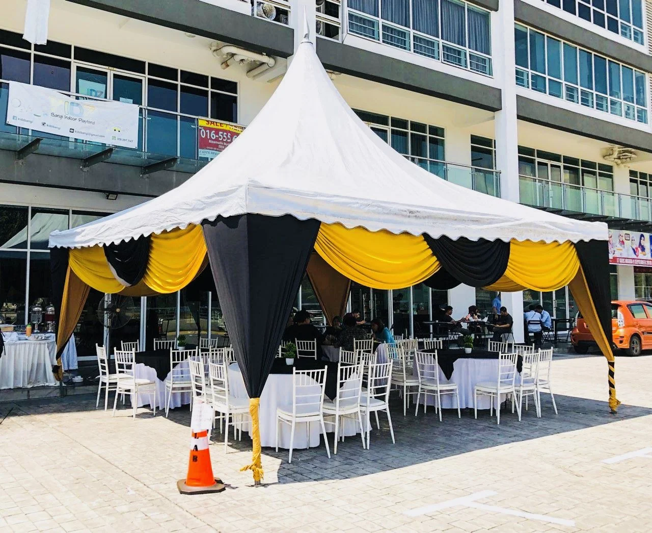 A large white event tent with black and yellow drapes, set up outside a modern building with glass balconies. Inside, there are tables covered with white and black tablecloths and white chairs. Some people are sitting and dining. An orange traffic co