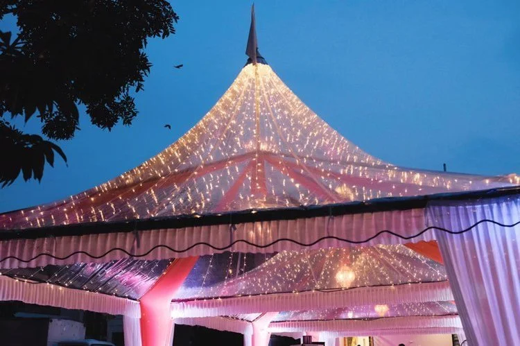 Decorated tent with string lights at dusk, with a flag on top and trees in the background.