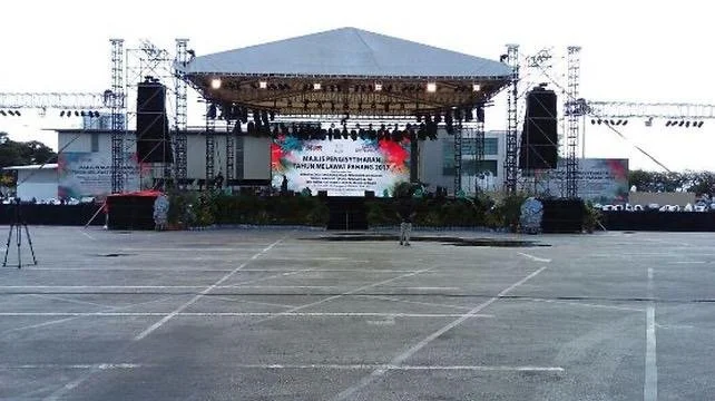 Empty outdoor stage with a large screen, speakers, and lighting setup, set up for an event, with a large open parking lot in front.