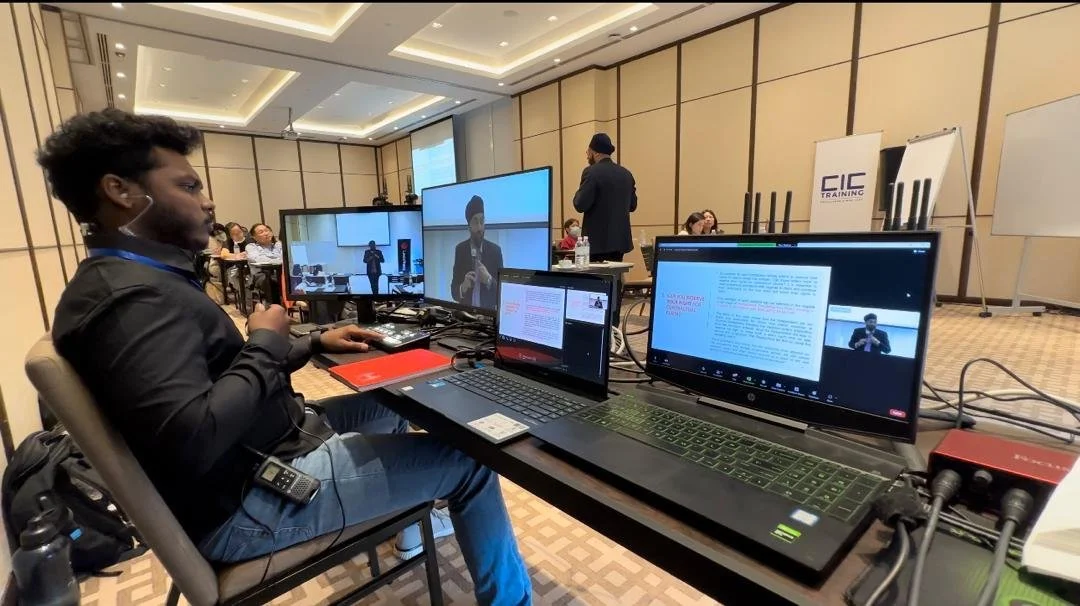 A conference room with several people, a man with a beard and headphones working on a laptop and multiple monitors, some screens showing a speaker and presentation slides, others with text and video calls, with a presenter standing at the front of th