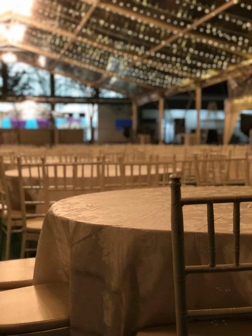 Empty outdoor dining area with wooden chairs and round table covered with a beige tablecloth, under a decorated ceiling with string lights, during evening.