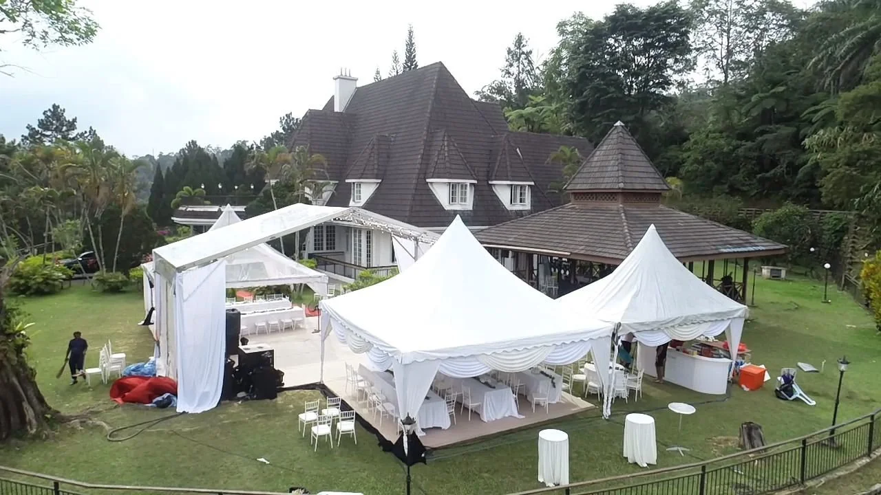 Outdoor setup with white tents, tables, and chairs in front of a large house on a well-manicured lawn, possibly for a wedding or event.