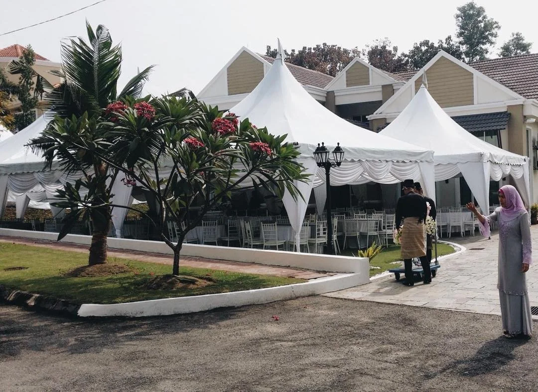 Large white event tents set up outside a building, with chairs and tables inside. There are two people outside; one standing and taking a photo or video with a smartphone, and another wearing traditional clothing, pushing a cart.