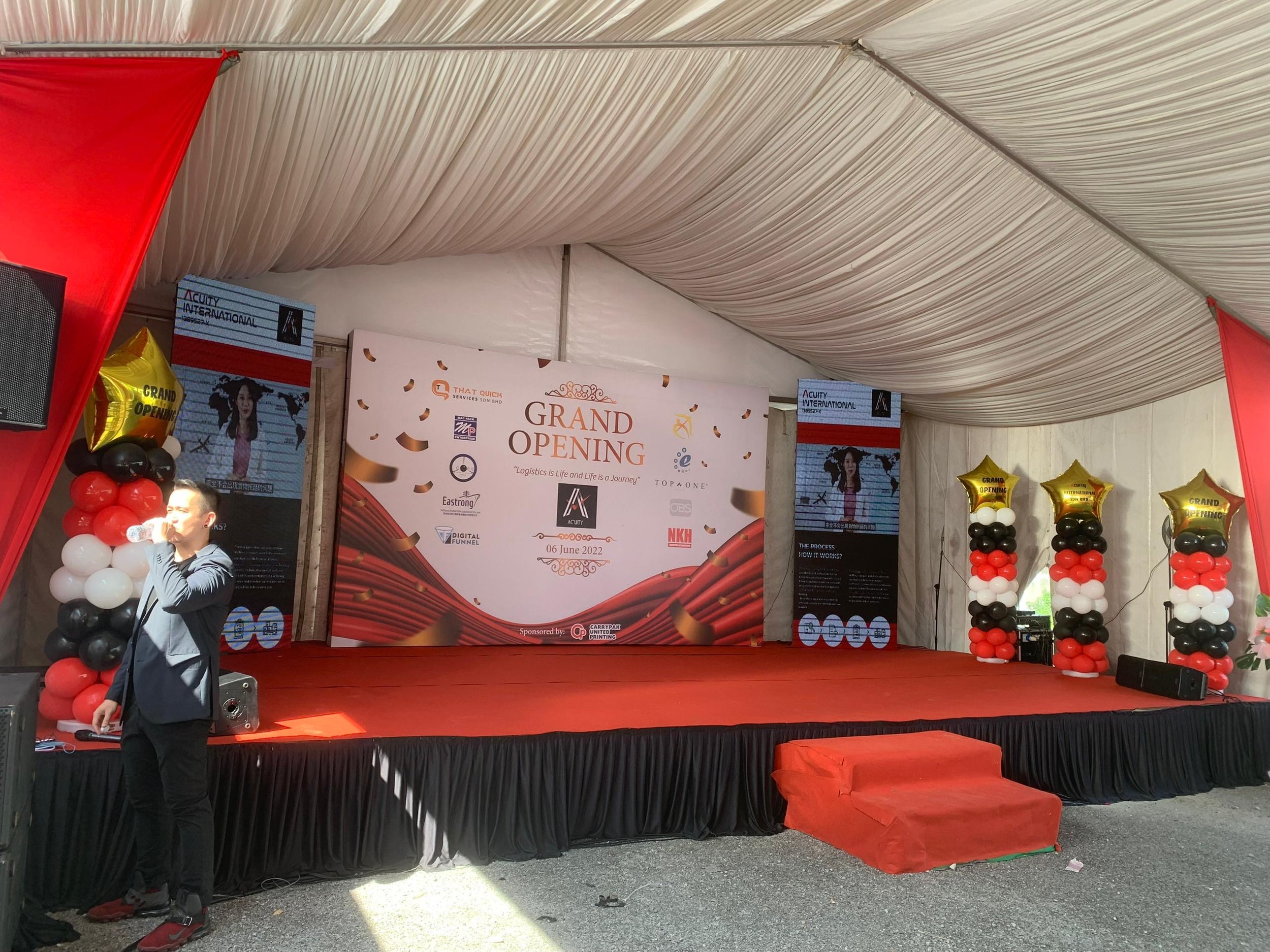 Event stage with a red and black balloon pillar, red carpet, and a large white backdrop with text celebrating a grand opening event.