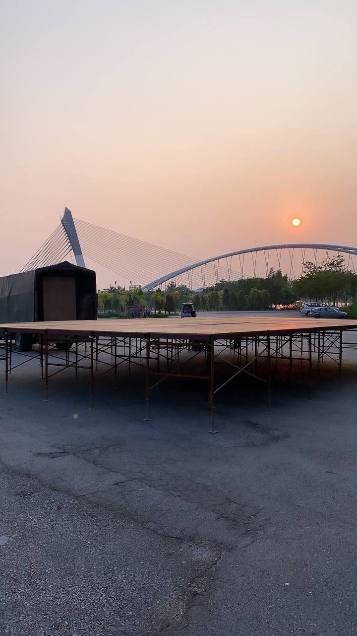 An empty outdoor stage with a backdrop of a modern arch bridge and a setting sun, surrounded by a parking lot and trees.