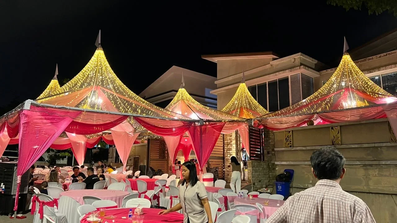 Outdoor event under a decorated tent with pink drapes, yellow string lights, and round tables with pink tablecloths, attended by people at night.