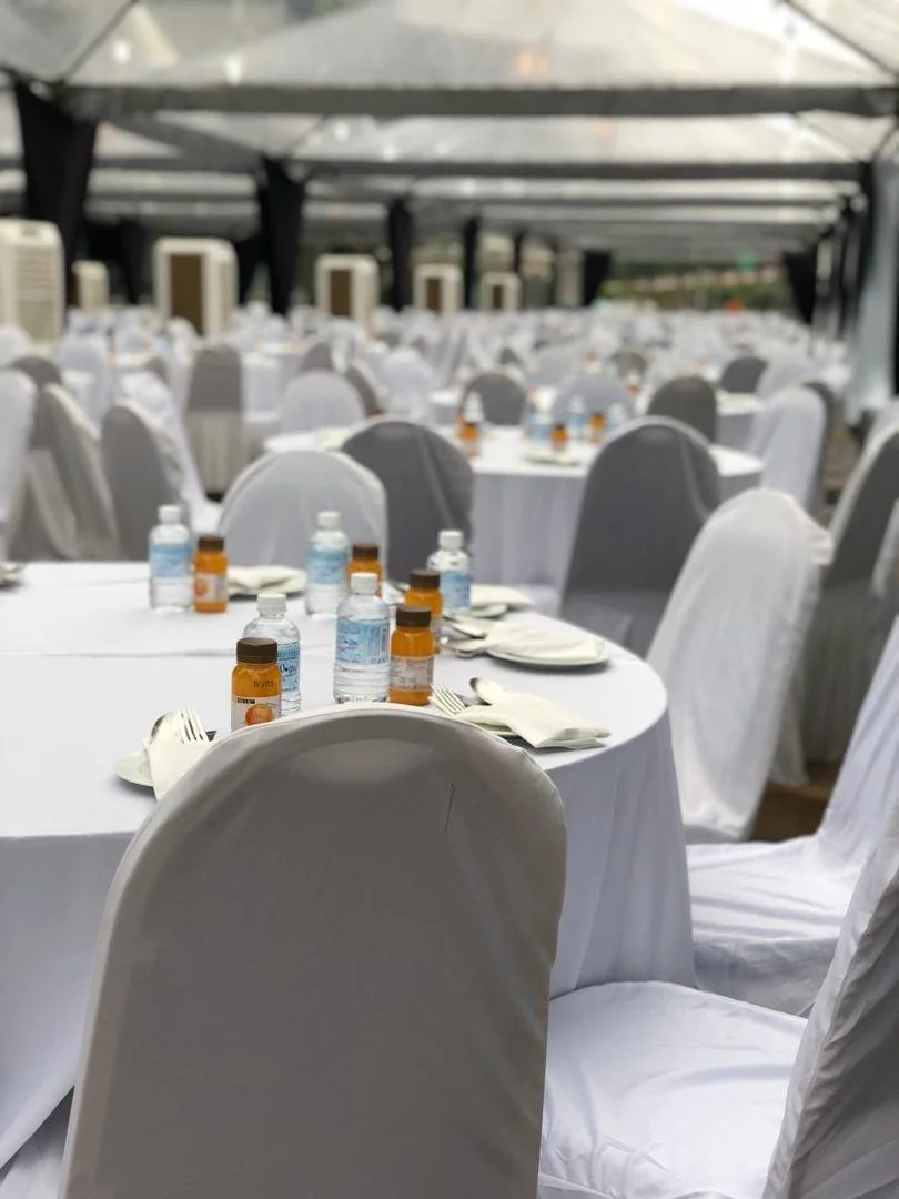 Empty banquet tables with white tablecloths, chairs covered with white slipcovers, bottled water and medicine bottles, set under a tent with a transparent roof.