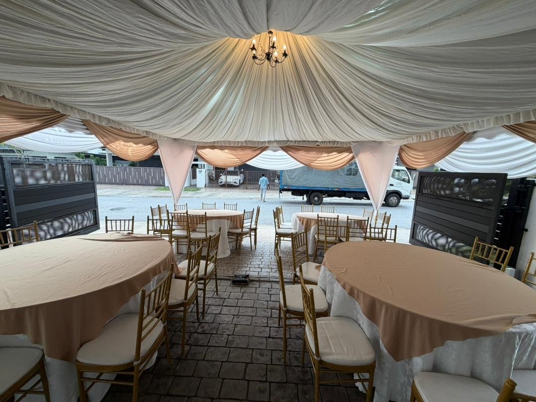 Indoor event space with draped fabric ceiling, chandelier, round tables with beige tablecloths, gold chairs, and an open view of the street outside with parked cars and a person walking.