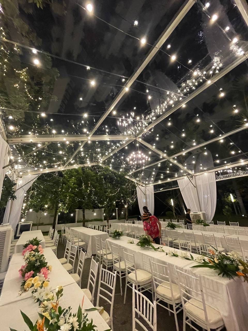 A decorated outdoor event space with string lights on a transparent ceiling, white draped curtains, long tables with white tablecloths, white chairs, and floral centerpieces. Two people are seen standing in the background, and trees with string light