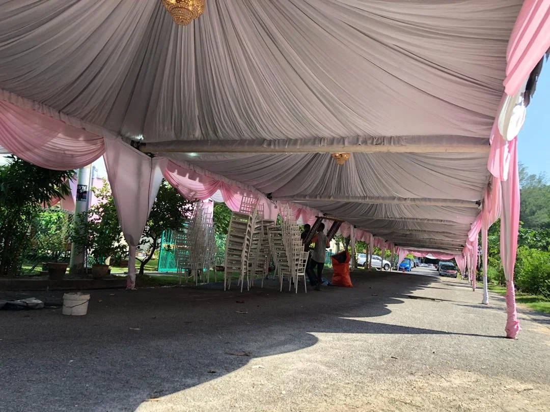 Large outdoor event tent decorated with pink and white drapes and a chandelier, with stacked chairs underneath and some people setting up.