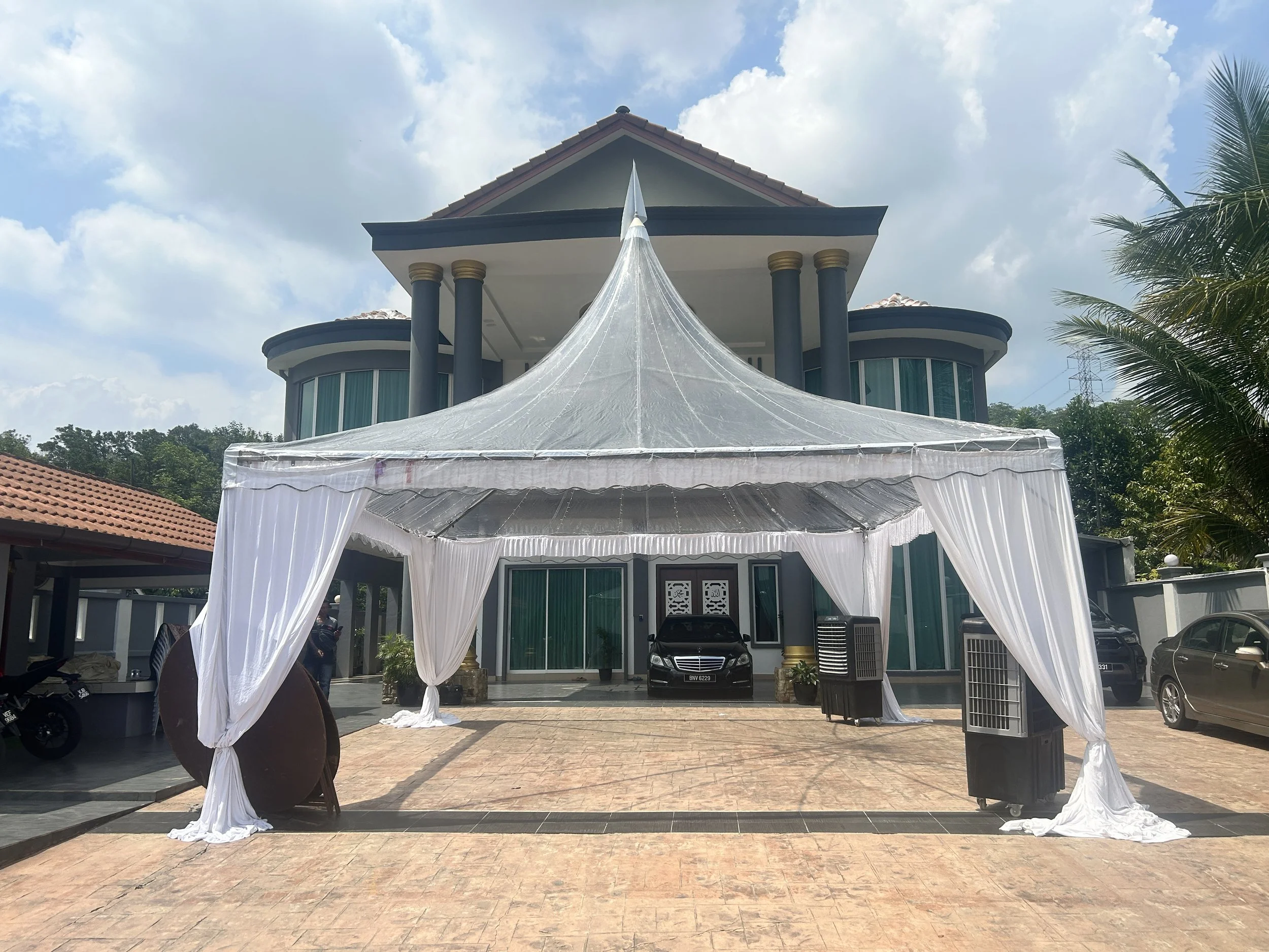 Large white event tent set up in front of a modern multi-story house with glass windows, with a black luxury car parked underneath, and palm trees to the right.