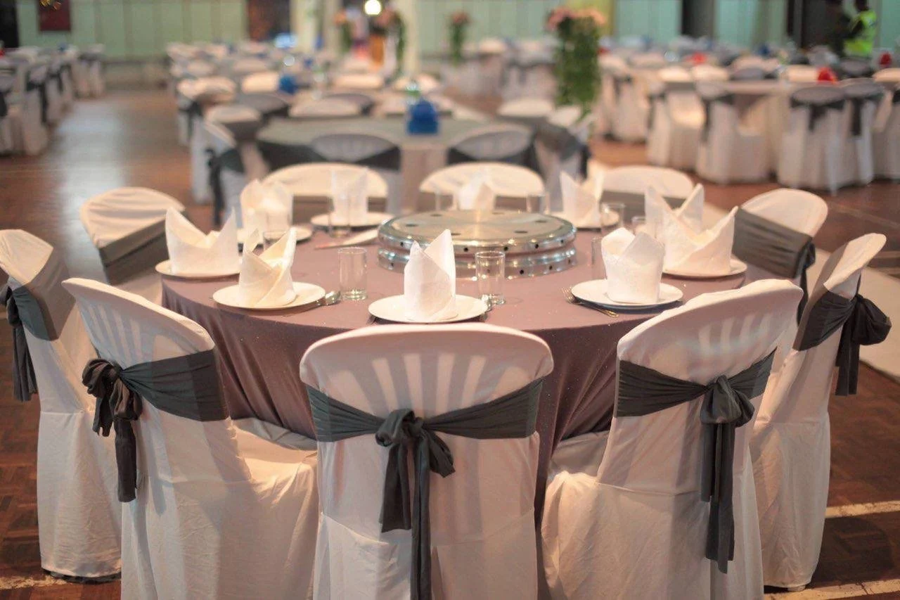 Round banquet table set for a formal event with white chairs tied with black sashes, white napkins folded into a standing shape, plates, glasses, and a central lazy Susan, in a decorated banquet hall.