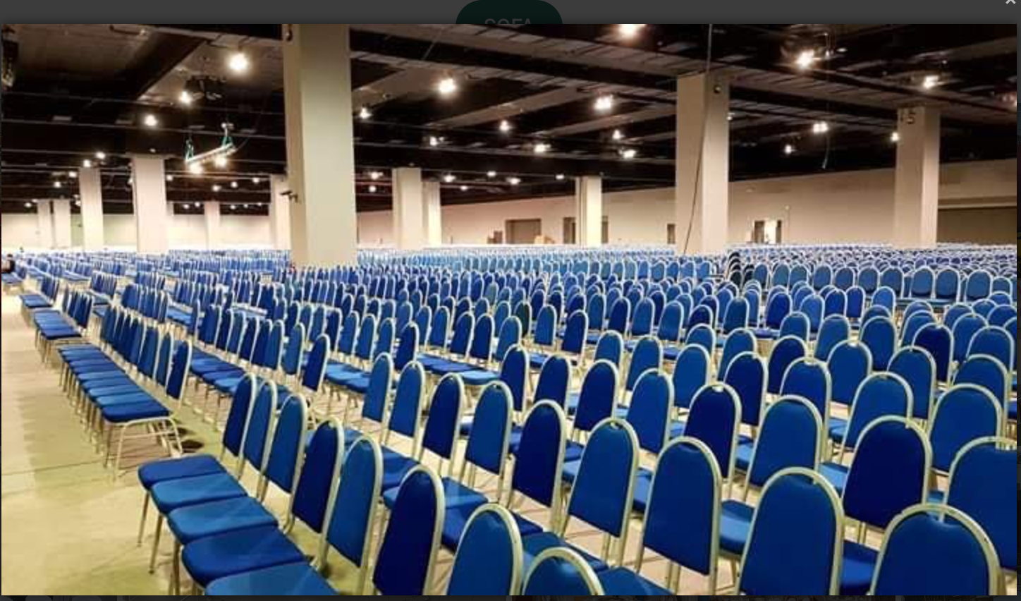Large hall filled with rows of empty blue chairs arranged in a grid, with some high ceiling lights and support columns.