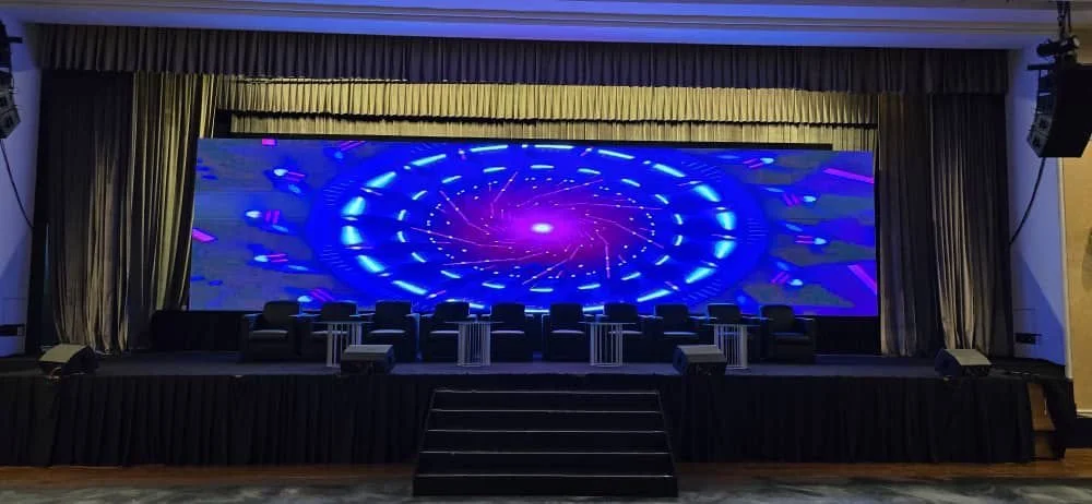 Stage with a large digital screen displaying a colorful, swirling digital graphic, surrounded by chairs and small tables, with curtains on either side.