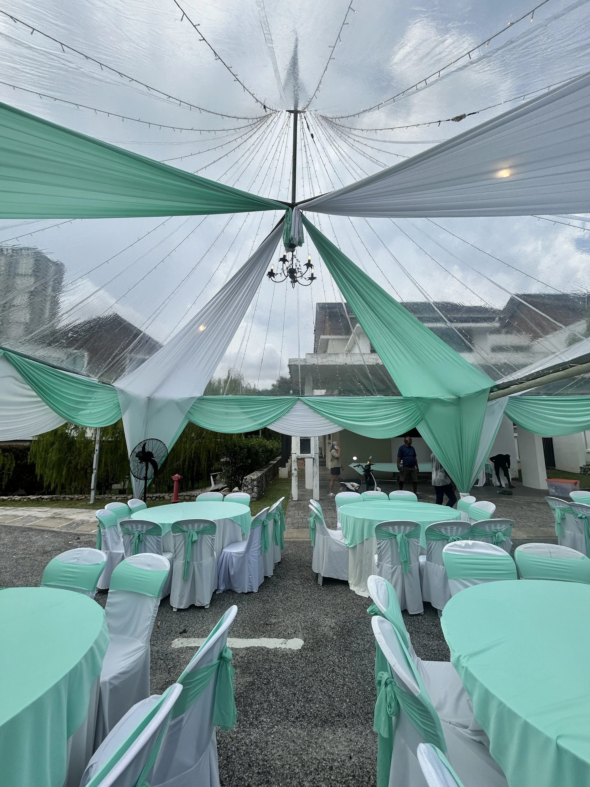 Outdoor event setup with round tables covered in white tablecloths and green table runners, and white chairs with green sashes. There is a decorative tent with green and white fabric draped underneath a transparent roof, and string lights hanging fro